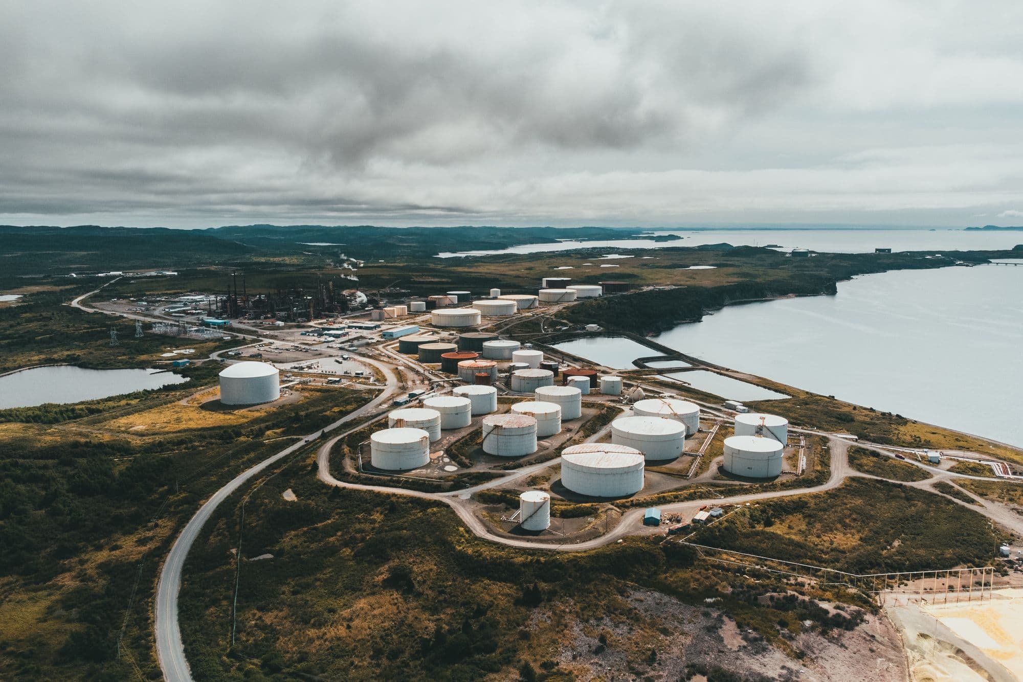 Aerial view of a coastal tank farm and industrial site beside a bay under heavy cloud